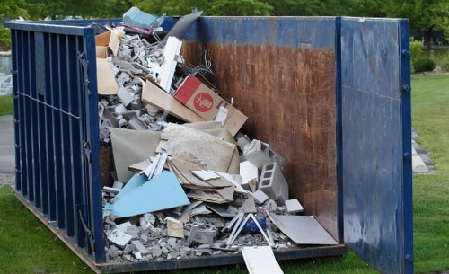 A large metal skip container placed outdoors on a grassy area, filled with a mixture of construction and household waste including broken drywall, cardboard boxes, plastic fragments, metallic scraps, and miscellaneous debris. The skip has a blue exterior with visible rust and uneven weathering, and its interior walls are brown from corrosion, contrasting with the white and grey waste materials. The waste appears to be loosely piled, spilling slightly over the edges of the container. In the background, green trees and a well-maintained lawn suggest a suburban or park environment, with natural daylight illuminating the scene. The image relates to rubbish removal services provided by Garden Clearance Islington, demonstrating the typical waste collected during garden or property clearance within the Islington area, postcode N1.