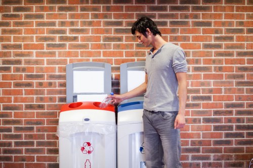 A young man with dark hair, wearing a light grey sweater and grey trousers, stands outdoors on a brick-paved area near a brick wall. He is holding a plastic bottle in his right hand, poised to dispose of it into a white recycling bin with a red recycling symbol on the front. Adjacent to the recycling bin is another similar bin with a blue lid for general rubbish disposal. The scene suggests a setting related to waste sorting and rubbish removal services in the area, possibly in Islington, given the context of the website, Garden Clearance Islington. The lighting appears natural, illuminating the scene evenly. The overall environment indicates an urban or residential outdoor space dedicated to waste collection and recycling efforts, aligned with professional rubbish removal practices.