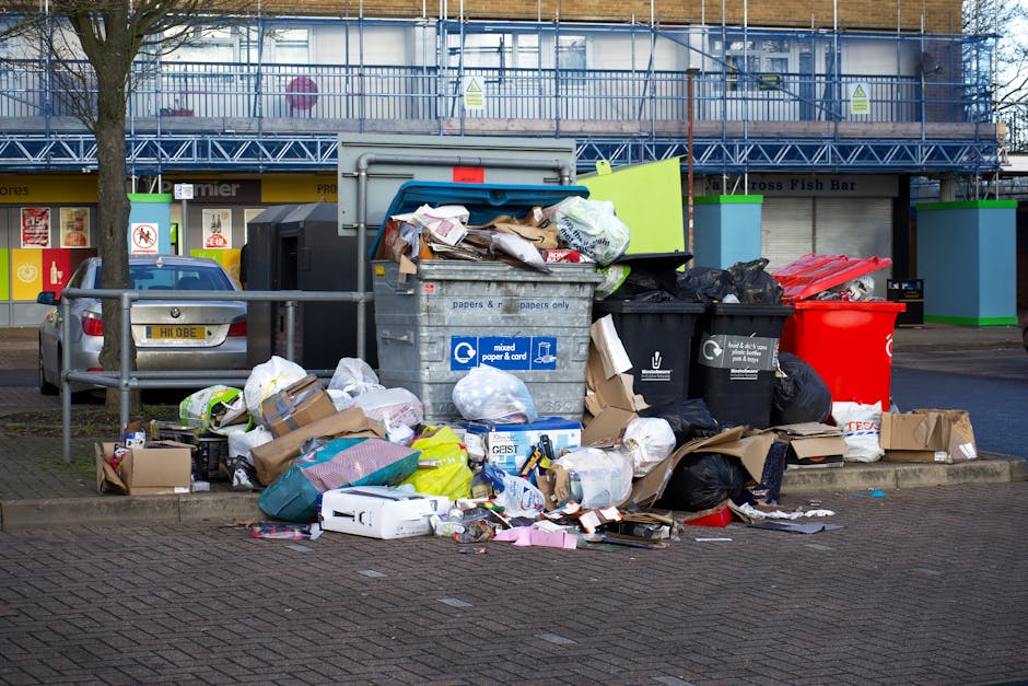 A large collection of mixed waste materials, including cardboard boxes, paper bags, plastic bags, and various discarded packaging, piled haphazardly on a paved urban sidewalk in front of multiple refu